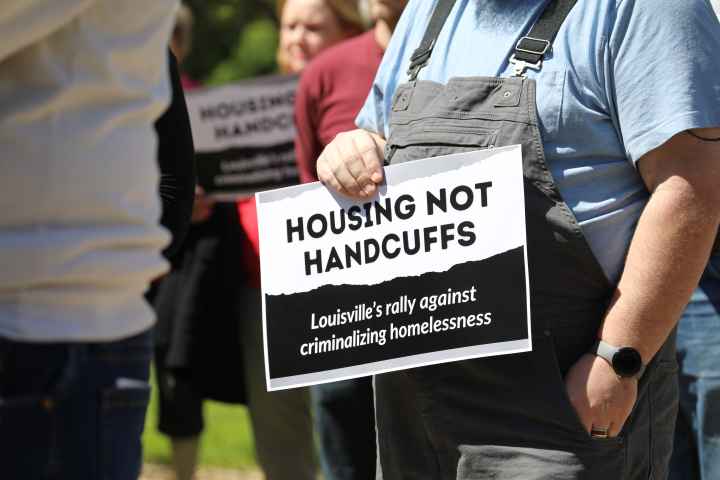 A person holds a sign that says "Housing Not Handcuffs, Louisville's Rally Against Criminalizing Houselessness"